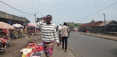 The image displays a daytime street scene in Goma, Democratic Republic of the Congo, characterized by an overcast sky. In the foreground, slightly left of center, a young woman walks forward on a concrete sidewalk. She wears a white long-sleeved shirt with black horizontal stripes and a vibrant, multi-colored patterned skirt. Her left hand is raised to her head. Immediately to her left, a market area is present on a dirt ground, featuring assorted garments in reds, whites, and yellows laid directly on the surface. Behind this, several vendor stalls operate under large umbrellas, including an orange one with an "Airtel" logo, displaying various goods and produce.

An asphalt road occupies the center and right portions of the image, extending into the background. Two men are observed walking away from the viewer on the road behind the central woman; one wears a light-colored top and dark trousers, carrying a white bag over his shoulder. A motorcycle is partially visible behind them. Along the left side of the road, a row of multi-story, unfinished concrete buildings with exposed rebar suggests commercial or residential use, with clothing hung from some structures. On the opposite side of the road, several single-story buildings with corrugated iron roofs are visible, one featuring a distinct stone or rubble wall. Overhead power lines traverse the sky, and a black modern streetlamp stands on the right side of the road. The scene portrays a bustling urban thoroughfare.