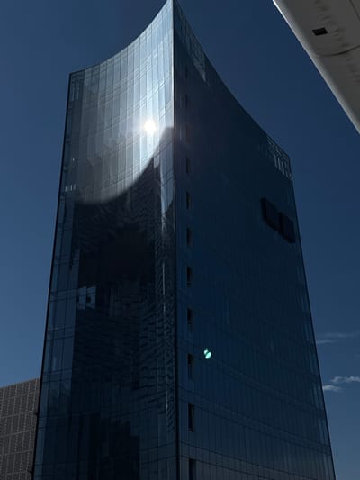 The low-angle image showcases a modern glass building against a clear blue sky. The building's architecture features a curved facade reflecting the sunlight, creating a bright glare. The rest of the building is dark blue with rectangular windows. A white object is at the upper left edge, possibly a part of another structure or vehicle. The architecture style is sleek and contemporary.