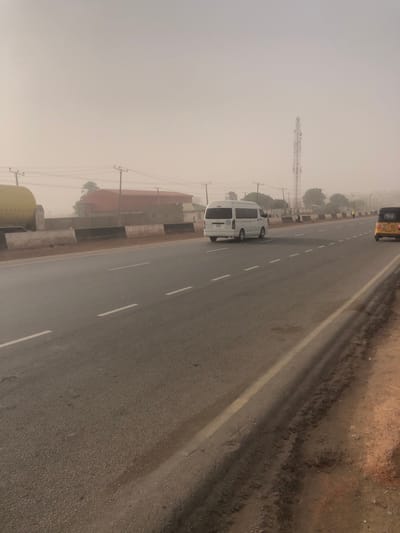 A hazy road view features a white van traveling on the left lane. A yellow vehicle is partially visible in the top right corner. Power lines run parallel to the road. A cell tower stands in the distance on the left. Adjacent to the road are buildings with red roofs, partially obscured by the haze. The sky is overcast, creating a muted color palette. Road markings are visible, indicating lane divisions.