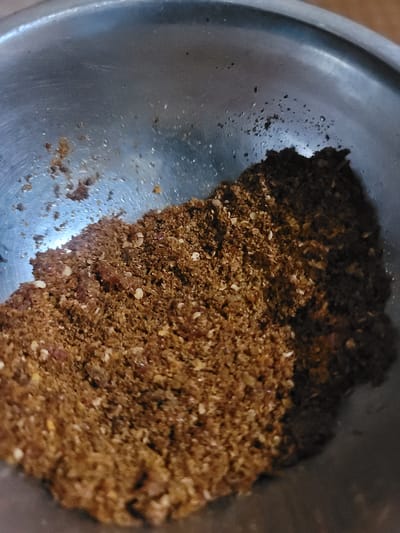 A close-up, overhead shot captures a stainless steel bowl filled with a mixture of finely ground spices. The spices are a rich, earthy brown color with flecks of lighter beige and some reddish-brown particles. The texture appears granular and somewhat clumpy. The bowl's interior shows some subtle reflections and a few water droplets, indicating it may have been recently washed or is part of a preparation process. The background is the interior of the bowl, which transitions into darker, out-of-focus areas. The image does not depict any specific location in Barakin Akawo, Nigeria, or any people or actions.
