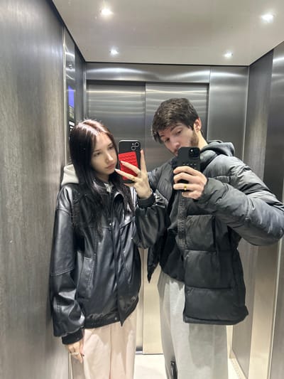 The scene depicts a mirror selfie taken by two individuals inside an elevator, located in Cachan, France.
On the left, a female individual with long, dark brown hair and a central part is visible. She wears a black faux-leather bomber jacket over a light-colored hooded top and light-colored trousers. Her right hand, adorned with red nail polish and multiple gold-colored rings, holds a black smartphone vertically, featuring a red, textured rectangular pop socket on its back, angled towards the mirror.
On the right, a male individual with short, dark brown hair and a stubble beard is present. He wears a black puffer jacket over a dark hooded top and light grey sweatpants. His right hand, displaying a gold-colored ring on the ring finger, holds a second black smartphone vertically, also angled towards the mirror.
Both individuals are looking in the direction of the camera capturing their reflection. The elevator interior features dark, horizontally paneled walls on the sides and reflective brushed metal doors at the rear. Two recessed ceiling lights are visible in the upper portion of the image.