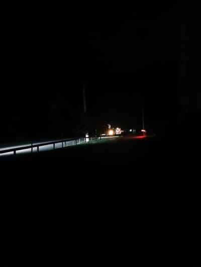 The image depicts a nighttime scene on what appears to be a road or highway. A light-colored guardrail runs horizontally across the lower portion of the frame, illuminated by a bright, white light source. To the right of the guardrail, the ground is dark and grassy. In the distance, there are faint lights indicating vehicles or structures. A bright red light is visible in the far distance, possibly taillights of a vehicle. A tall pole, possibly a utility pole, is also present in the background. The scene is dark, with limited visibility beyond the immediate area illuminated by the guardrail lights and distant lights. There is no indication of the specific location within Poltava, Ukraine.