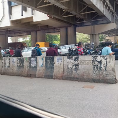 This photograph captures an urban scene in Bengaluru, India, likely under a flyover or metro bridge. The setting is a busy road, with vehicles and pedestrians appearing to be stopped or moving slowly in traffic.

In the foreground, a row of concrete traffic barriers separates the viewer's perspective from the main road. These barriers are heavily stained and defaced with peeling posters and graffiti. Several men are visible behind these barriers, some standing, some on motorbikes, and one leaning against a barrier while looking at his phone. They appear to be waiting in traffic. Cars and motorbikes are queued behind them, and in the distance, a yellow truck and another blue vehicle are visible.

Above the street, a massive concrete and metal structure forms an overpass, possibly a metro line or an elevated road. The intricate metal framework and large concrete pillars supporting this structure are prominent.

Notable details include the daytime lighting, suggesting a bright but possibly overcast or hazy day. The overall impression is one of a typical bustling Indian city street.

Visible text on the concrete barriers includes:
*   Repeated phrases related to "AI GEN", "AGENTIC", "AI AGENTS".
*   "BOFFUM CLASSES" and a phone number "88 499 495".
*   "TECHNOLOGY", "TRAINING WITH REAL TIME PROJECTS", "GENERATIVE AI", "AGENTIC AI LLM".
*   In Kannada script, "ಬಿಬಿಎಂಪಿ" (BBMP), which stands for Bruhat Bengaluru Mahanagara Palike, the municipal corporation of Bengaluru.
A car license plate "KA01LT458" is also visible, confirming the location as Karnataka.