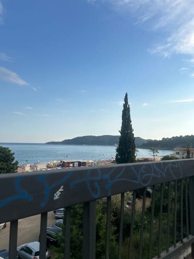 The image shows a view from an elevated position overlooking a beach and the Adriatic Sea. In the foreground, a dark metal railing with blue graffiti is visible. Behind the railing, a parking area with several cars is present, along with lush green vegetation.

Beyond the parking area, a busy beach scene unfolds. Numerous people are seen relaxing on the sand, swimming in the water, and sunbathing. Beach umbrellas, lounge chairs, and small structures, possibly changing rooms or snack bars, are set up along the shoreline. The sea is calm and blue, with some gentle waves reaching the shore.

In the middle ground, a tall, slender cypress tree stands prominently. The background features rolling hills covered in dense green vegetation, indicative of a coastal landscape. A few buildings are nestled along the coastline, and a large white yacht is visible on the water in the distance. The sky is a clear, bright blue with a few wispy white clouds, suggesting a sunny day.

This scene is consistent with the coastal town of Boreti, Montenegro, known for its beaches and scenic views of the Adriatic Sea and surrounding hills.