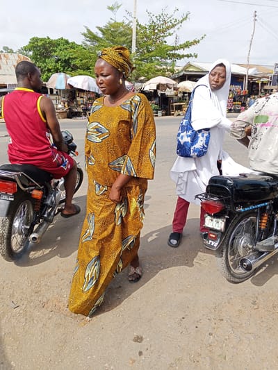 The photo was taken in Zango-ndaji, Nigeria. It depicts a woman in a mustard yellow dress and headwrap walking past two motorcycles. One motorcycle has a man in a red shirt and shorts sitting on it. The other motorcycle has a woman in a white hijab standing next to it and a man in a white sweater partially in the frame. In the background, there are several stalls with umbrellas covering them. Trees and buildings are visible behind the stalls. The ground is a light-colored pavement. The sky is partly cloudy.