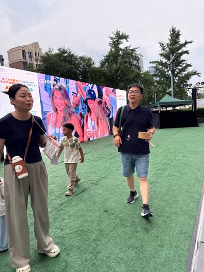 The image depicts an outdoor scene with several people standing on artificial turf in front of a large digital billboard. The billboard displays vibrant images of people, some with tattoos, and features Chinese text. The text on the billboard includes "第二十四届中关村国际美食节", "暨清河街道", "米·味来已来", "中共北京海淀区清河街道工作委员会 办事处", and "AI農食嘉年华". A logo for "CCTV 5 央视频" is also visible.
In the foreground, a woman in a dark top and grey pants is seated, looking towards the right. She is carrying a white thermos with a brown leather case and a cartoon design. To her right, a young boy in a light-colored t-shirt with a tree print is standing, holding a folded paper. Behind the boy, a man in a dark polo shirt and denim shorts is standing and holding a small wooden object. He is wearing glasses. The overall atmosphere appears casual and possibly related to a festival or promotional event, given the billboard and the presence of food-related text. The sky is overcast, suggesting a cloudy day. Buildings and trees are visible in the background.