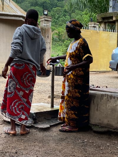 This image captures an outdoor scene in Mararaba, Nigeria, depicting two individuals at a water collection point. The ground is unpaved, composed of damp, light brown dirt and gravel.

On the left, an individual is observed from the back, wearing a grey hooded sweatshirt and a long, red skirt with intricate white and grey spiral and geometric patterns. Their hair features dark braids with reddish accents visible beneath the hood. They wear flat, brown sandals with metallic straps and hold a dark blue bowl in their left hand, positioned near the water tap.

On the right, an individual is shown in profile, facing left, adorned with a patterned headwrap and a long, loose-fitting dress (kaftan) displaying white floral designs amidst speckled brown, black, and yellow patterns. They wear black flip-flops with pink straps. This individual is actively operating a silver metallic water tap with their right hand, while holding a silver metallic pot beneath the flowing water with their left hand.

The water source is a metal tap extending from a vertical pipe embedded in the ground. In the background, between the two individuals, a concrete pillar supports a black floodlight. To the right, a yellow metal gate with decorative spikes is partially visible, behind which a light blue car is parked. A portion of a building with a light-colored roof is seen on the far left. The distant background is characterized by a densely forested hillside, suggesting a lush environment under an overcast sky. In the immediate foreground on the right, a low concrete structure, possibly a well cover, with a wooden top is present.