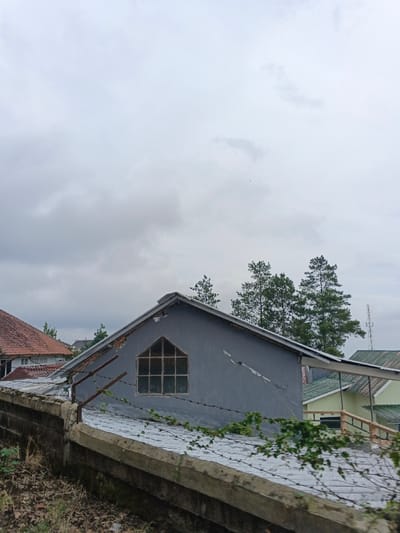 The image displays a residential scene in Kabiufa, Papua New Guinea, under a heavily overcast sky. In the foreground, a concrete and brick retaining wall runs horizontally, topped with multiple strands of rusted barbed wire. Green vine-like vegetation is intertwined with the barbed wire. Behind this wall, a two-story building with a grey-painted gable end is prominent, featuring a distinct arch-topped, multi-paned window. Diagonal cracks are visible on the grey facade of this building. Its corrugated metal roof is partially visible behind the wall. To the left, a building with a reddish-brown tiled roof is partially obscured. To the right and further back, other structures with light green and white roofs are present. Several tall evergreen trees rise in the background, and the upper section of a metallic communication tower is visible in the far right distance. No individuals or discernible activities are present.