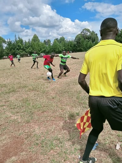 The image depicts an outdoor soccer game in Lwandeti ward, Kenya, under a blue sky with scattered white cumulus clouds. In the immediate foreground on the right, the back of a male individual is visible, dressed in a bright yellow short-sleeved shirt with black trim, black shorts, and black socks, holding a red and yellow checkered flag, consistent with an assistant referee. In the midground, five male individuals are actively participating in the game on a dry, grassy field with patches of bare earth. A central player, wearing a red short-sleeved shirt and green shorts, dribbles a white soccer ball with black pentagons. To his immediate right, another player in a green and black striped short-sleeved shirt with "pepets" branding and black shorts moves towards him, arms outstretched. Further back, another player in a similar green and black striped uniform runs to the left. Two additional players in green and black striped uniforms are visible in the left background, near what appears to be a white goalpost frame. A dense line of green trees forms the distant background horizon. All visible players are male.