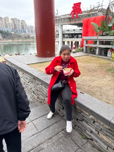 The image presents an outdoor scene, likely a riverfront promenade or park, situated in what appears to be an urban area consistent with the Shapingba District of Chongqing, China. The sky is overcast.

In the mid-ground, an elderly Asian woman with short, dark hair streaked with grey is seated on a low, grey stone wall. She wears a vibrant red long coat with prominent white, circular buttons, a patterned dark red and purple sweater, dark grey trousers, and white athletic shoes with red accents. She is holding a white paper cup, adorned with red Chinese characters, in her left hand, and a spoon or small fork in her right hand, positioned near her mouth as if consuming food. A black shoulder bag rests on her left hip. Her expression is direct, looking towards the viewer. Several small, dark-red, irregular objects, possibly nuts or seeds, are visible on the stone wall next to her.

In the lower-left foreground, the back and partial left side of an individual wearing a black jacket and dark trousers are visible, facing away from the camera. The ground beneath consists of segmented grey stone paving, featuring visible seams and a change in elevation suggesting steps.

The background features a wide river. Across the river, a dense skyline of numerous tall, modern buildings rises along a hillside. To the left of the seated woman, a substantial, dark red cylindrical column extends vertically. Behind her, a multi-level bridge structure is visible. Above this bridge, a large, red, inflated or fabric depiction of the Chinese character "Fu" (福) is prominently displayed. To the right of this character, another large, red, inflatable structure is partially obscured by foliage. A red and white striped traffic cone is also present in the background near these decorative elements. The area directly behind the stone wall is a sparse, light brown grassy patch.