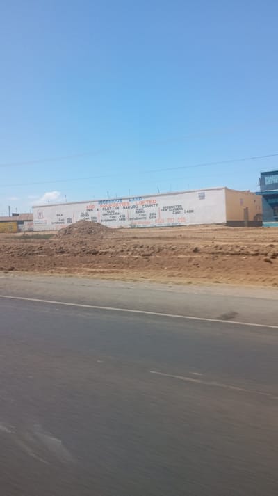 A roadside advertisement for Incenious Land & Properties Limited in Nakuru County, Kenya. The ad promotes land ownership, listing property areas like "Sola", "LancaLanga", "Ilave Sooro", and "Greensted View Gardens" along with cash and installment prices. A phone number, "0723 777 555", is displayed prominently. The advertisement is painted on a long white building next to a brown dirt area. The blue sky is visible in the background.