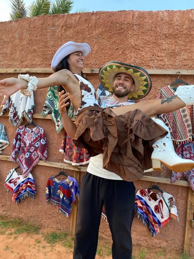 A man in a sombrero and patterned shirt is holding a woman in a cowboy hat and cow-print top. The woman is wearing a brown ruffled skirt and white cowboy boots, and her legs are extended. They are posing in front of a wall of colorful woven ponchos hanging on wooden racks. The wall appears to be made of reddish-brown stucco. Palm fronds are visible at the top of the image, suggesting an outdoor location with a warm climate. The ground in the foreground is sandy and has some sparse green vegetation. The scene has a festive, possibly tourist-oriented atmosphere, and the attire suggests a theme of Western or Mexican culture. Given the context, the location is likely a market or a stall selling souvenirs in Mui Ne, Vietnam.