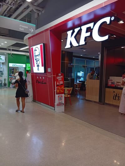 The image shows the exterior of a KFC restaurant. The KFC logo is prominently displayed in white letters on a red background above the entrance. To the left of the KFC, there is a green and white sign for "CURRENCY EXCHANGE." A woman in a black dress is walking away from the camera towards the left of the frame, carrying a small purse. In the background, several people are visible inside the currency exchange and the KFC restaurant. The floor is tiled, and the ceiling has visible pipes and lighting fixtures. A sign pointing to the right with "Exit" written on it is also visible on the left side of the frame. The overall environment appears to be an indoor mall or shopping center.
