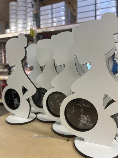 The image shows a stack of white, cutout frames in the shape of a pregnant woman. Each frame has a circular opening where an ultrasound image is displayed. The ultrasound images show fetuses at various stages of development. The frames are layered on top of each other, creating a sense of depth. The background is blurred but appears to be an indoor setting, possibly a store or a workshop, with shelves and other objects visible. The overall impression is one of anticipation and celebration of new life. The frames are made of a matte material, possibly wood or thick cardstock, with a black border around the circular openings. Some of the frames have a slight sheen, suggesting they may be laminated. The lighting in the image is soft and even, with no harsh shadows. The focus is on the frames and the ultrasound images, drawing the viewer's attention to the delicate details of the developing fetuses. The scene evokes a sense of tenderness and wonder.