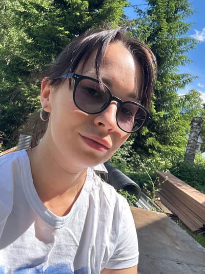 In Novozhilovo, Russia, a young woman with dark hair, wearing a white t-shirt and black sunglasses, is taking a selfie outdoors on a sunny day. She is positioned in the foreground, with lush green trees and a bright blue sky with scattered clouds forming the background. To the right, a stack of wooden planks and some construction materials are visible, hinting at a possible construction or renovation site. The woman is looking towards the camera with a slight smile.