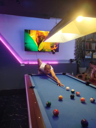 A man is playing billiards in a dimly lit establishment in Patong, Thailand. He is positioned at the blue felt-covered pool table, leaning over to strike a white cue ball with his cue stick. Several other billiard balls, some with visible numbers and colors, are scattered across the table. In the background, a large television screen displays an image, and neon pink lighting illuminates the wall. To the right, two individuals are seated, one of whom is smoking a cigarette. The floor is dark and reflective.
