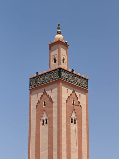 The image shows the minaret of the Koutoubia Mosque in Marrakesh, Morocco, viewed from a low angle. The minaret is a tall tower with alternating light and dark pink stripes. It is decorated with geometric patterns in green, blue, and gold tiles near the top. The top of the minaret is adorned with golden orbs. The sky is a clear blue.