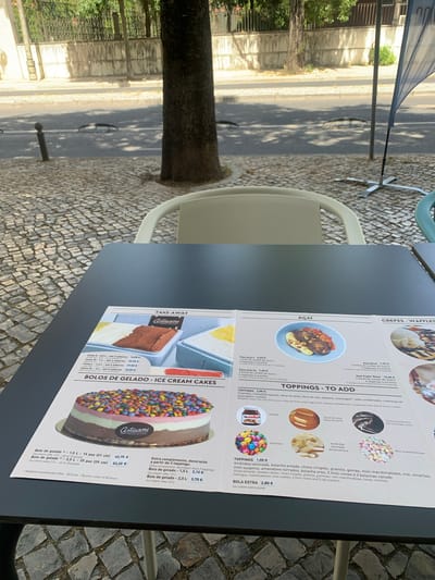 The image shows a close-up view of a laminated menu placed on a dark gray table. The menu is open to a section featuring desserts, with several appetizing pictures and descriptions. The menu items include "BOLOS DE GELADO - ICE CREAM CAKES," "AÇAÍ," and "CREPES - WAFFLES."
The "ICE CREAM CAKES" section displays a colorful, multi-layered cake adorned with small candies on top. Below this image, there are descriptions of cake sizes and prices, such as "Bolo de gelado - 1,5 L - 15 pax (21 cm)" priced at 42,75 €. The text also mentions "Meidante encomenda prévia - 48 horas / By prior order of 48 hours."
The "AÇAÍ" section shows a bowl of açai topped with fruit, granola, and other toppings, with options for different sizes and prices like "Taça Açaí L, 5,60 €."
Further to the right, the "CREPES - WAFFLES" section is partially visible, showing a waffle with ice cream and chocolate sauce.
Below these sections is a "TOPPINGS - TO ADD" section, with images and names of various toppings like "Nutella," "Doce de Leite," "Chocolate Quente," "Smarties," "Amêndoa laminada," and "Mini-marshmallows." There is also a "BOLA EXTRA 2,80 €" for an extra scoop.
The menu is printed in Portuguese with some English translations. The overall lighting suggests it's daytime, possibly outdoors, as indicated by the dappled sunlight on the pavement outside and the presence of a street and trees in the background. The pavement in the foreground is made of cobblestones. The establishment appears to be a cafe or restaurant in Lisbon, Portugal, specializing in ice cream and desserts. The outdoor setting, with the cobblestone street and trees, is characteristic of many areas in Lisbon.
