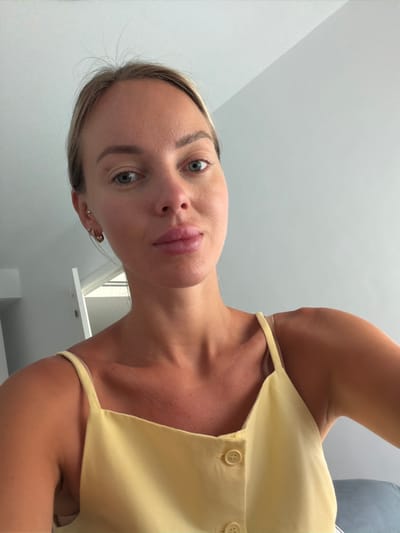 The image shows a close-up selfie of a young Caucasian woman. She has fair skin, light brown hair pulled back, and blue-green eyes. Her lips are full and painted a soft pink. She is wearing a pale yellow camisole with thin straps and two visible buttons. She has small earrings in her left ear. The background is a plain, light gray wall, suggesting an indoor setting. The lighting is bright, casting minimal shadows. The image does not contain any specific landmarks or elements that would indicate its location within Finestrat, Spain.