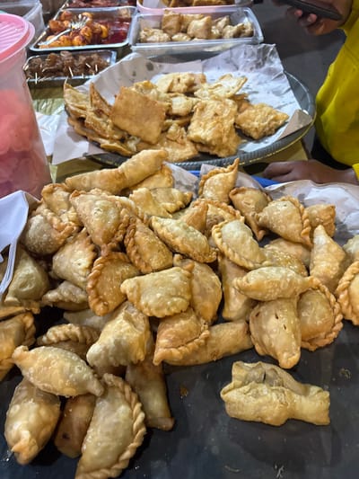 This image captures a close-up view of a food stall in North Jakarta, Indonesia, likely a night market or a street food vendor. The main focus is a large pile of freshly fried savory pastries, resembling curry puffs or empanadas, displayed on a dark surface. These pastries are golden brown and have a textured, crimped edge. To the left, a pink plastic container with a ridged lid is visible, hinting at a sauce or condiment. In the background, trays of other fried snacks and food items are arranged, some appearing to be skewered and coated in sauce. A person in a bright yellow hoodie is partially visible on the right side of the frame, holding a smartphone, suggesting a customer or vendor interacting with the food. The overall environment appears to be an outdoor food setting, typical of Indonesian street food scenes.