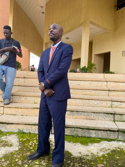 The media file depicts two individuals outdoors, possibly in Nigeria, given the location context. The main subject is a bald Black man wearing a navy blue suit, an orange tie, and black dress shoes. He is standing and looking to his left, with his hands clasped in front of him. He appears to be in his 40s or 50s and has a goatee. He has a serious expression. Behind him, a younger Black man is partially visible, wearing a navy blue t-shirt, blue jeans, and grey sneakers. He is holding a phone and a bag, and appears to be looking down.
The scene is set against a backdrop of a building with a yellow facade and a staircase made of large, textured concrete blocks. Moss and small plants are growing on the steps and the ground to the right, suggesting a humid or damp environment. The lighting suggests it is daytime, possibly overcast due to the diffused light. There is a small sign on the wall of the building with some text, but it is not legible in the image. The overall impression is that of a formal or semi-formal gathering, or perhaps a casual encounter.
