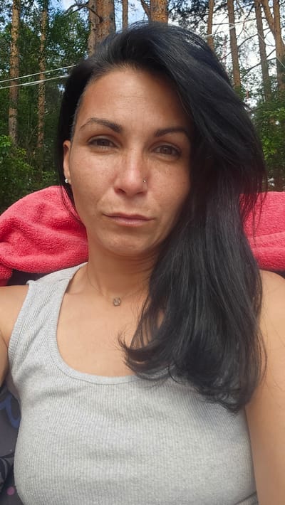 A woman with dark hair and a light gray tank top is pictured outdoors. She is looking towards the camera. Behind her, a red towel is visible, and beyond that, trees and foliage suggest a natural setting. The image appears to be taken in Dolni Pasarel, Bulgaria.