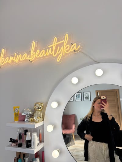 A woman is taking a selfie in a mirror. She is wearing a black tank top and a white sequined skirt. Her hair is blonde and she is wearing a necklace. In the mirror, we can see a room with a chair and a doorway. There is also a neon sign on the wall that says "karinabeautyka". On a shelf to the left of the mirror are several beauty products, including a tube of hand cream, a disco ball, and a gold teddy bear figurine. The lighting in the room is soft and warm. The setting appears to be a beauty salon or a home studio. It is not possible to determine the city of Riga, Latvia from the image.