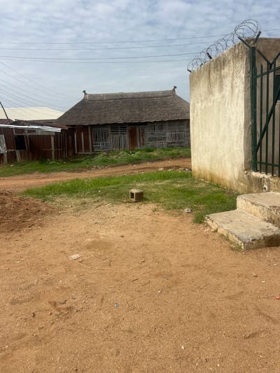 The image depicts an outdoor scene with a traditional-style building featuring a thatched roof and walls constructed from horizontal wooden slats, located in the city of Rafin Par, Nigeria. Adjacent to this building are structures with corrugated metal roofing and siding. In the foreground, a dirt path leads past a pile of soil and patches of grass. To the right, a concrete wall topped with barbed wire surrounds a metal gate, and a set of concrete steps descends from this area. The environment appears to be an urban or semi-urban setting, with power lines visible against a cloudy sky. No people are present in the image, and no specific actions or interactions are depicted. The overall scene is one of everyday life in a developing area.
