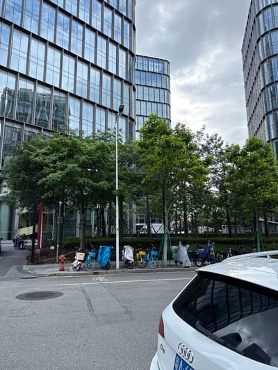 A street scene in Haizhu District, China, features modern glass-fronted office buildings and a line of parked electric scooters and bicycles. The buildings have a contemporary architectural style with repeating window panes that reflect the overcast sky. Trees with lush green foliage are planted along the sidewalk, interspersed with streetlights and what appears to be construction scaffolding. In the foreground, a white SUV is parked on the asphalt road, partially obscuring the view of the sidewalk. A red fire hydrant is visible near the curb, along with several electric scooters covered with protective tarps and two blue bicycles. A few individuals are seen in the background, near a building entrance, appearing to push carts. The street is marked with painted lines indicating traffic lanes and a bicycle lane. The overall environment suggests an urban setting with a blend of modern infrastructure and green spaces.