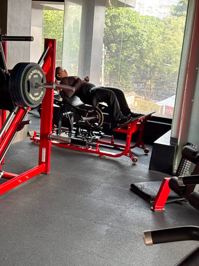 A woman is using a red leg press machine at a gym in Kolkata, India. The woman is wearing a brown jacket and black pants and is lying at a 45-degree angle. Weights are stacked on the foot platform. A window behind her shows trees and buildings outside. Gray rubber gym flooring is visible. Another piece of gym equipment is visible in the upper right.