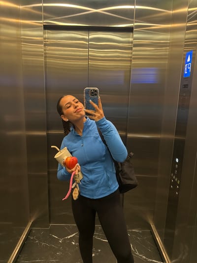 A woman stands in an elevator with stainless steel walls, holding a smartphone in her left hand to take a selfie. In her right hand, she holds a red apple, a white reusable coffee cup, and a keychain with keys attached to a pink woven strap. She is wearing a blue athletic jacket, black leggings, and has a black backpack slung over her shoulder. The elevator has a control panel with illuminated floor numbers and call buttons. The floor of the elevator is dark marble with white veining. The location is not indicated by visual cues in the image.