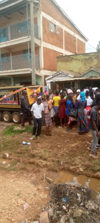 The scene takes place in a small town in Kivaywa, Kenya. A group of approximately 30 people are gathered around a hearse. The hearse is decorated with yellow and gold fabric.  It is parked in front of a brick building that has a sign for "MATETE SALON & COSMETICS" above the entrance. The people are dressed in a variety of casual clothing and are talking to each other.  Some of them are holding flowers and are walking behind the hearse. The hearse is facing a dirt road.  The road is filled with garbage and is lined by a small, shallow ditch filled with murky water.  The ground is dirt and grass. The sky is partly cloudy.