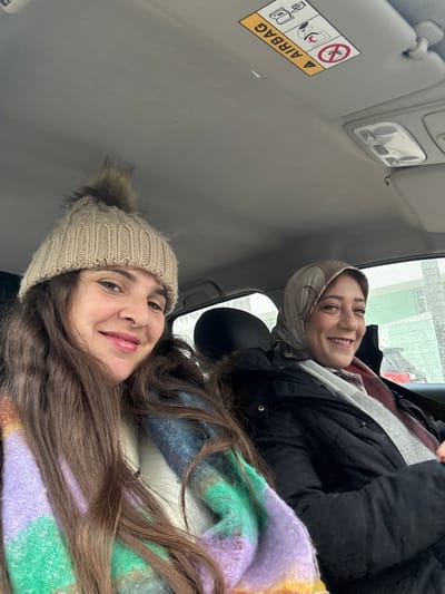 The image displays two adult females inside a vehicle, likely a car, captured in a selfie. The vehicle's interior features a grey headliner, a sun visor on the passenger side with a yellow "AIRBAG" warning label, and an overhead console light fixture. The individual on the left, positioned in the foreground, has long brown hair and wears a light beige ribbed knitted beanie with a brown faux fur pom-pom. Her lips are glossy pink, and she is smiling directly at the camera. She wears a multi-colored chunky knit scarf with prominent bands of purple, green, and blue, over a white garment. The individual on the right, positioned in the background, wears a light brown patterned headscarf (hijab), a black quilted jacket with a fur-lined collar, and a grey and reddish-patterned scarf. She is also smiling towards the camera. Through the vehicle's window, an overcast or rainy exterior is suggested by blurred reflections, with a red vehicle partially visible in the distance. While the image is associated with Meknes, Morocco, no specific landmarks are visible to pinpoint the exact location within the city.