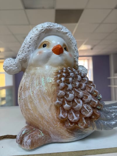 A close-up, slightly out-of-focus image shows a decorative, avian-themed ceramic figurine. The figurine has a white head with a textured, hood-like feature. It has an orange beak, and its body is brownish with a scale-like texture on the top, sprinkled with glitter. The background includes a wall with square, white tiles and a purple wall section. The location is not identifiable as Klin, Russia from the provided image.