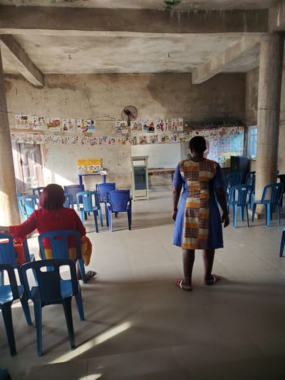 The media file captures an indoor scene, likely a classroom or learning center, in Ukwu-Abwa, Nigeria. The room features concrete walls and floor, with exposed concrete beams in the ceiling, giving it a somewhat utilitarian appearance.
Two individuals are visible. In the foreground, a child, viewed from behind, is seated in a blue plastic chair, wearing a red top. Further back, an adult stands, also viewed from behind, wearing a blue dress with a distinct yellow and dark patterned mid-section, and pink and white sandals.
The room is furnished with numerous blue plastic chairs, some arranged in what appears to be rows, and others stacked. Bright natural light streams in from a window, creating strong sunbeams on the floor.
A prominent feature is a series of educational posters or charts strung along one wall with clothespins. These charts display letters of the alphabet and corresponding words or images, indicative of early childhood education materials. Visible text includes "Y y", "X X N", "ch", "yogurt", "x-ray choo-chao", "H h", "thumb", "QU", "quack", and multiple instances of "M M M M", "K", and "M". Another large chart, possibly an alphabet or vocabulary chart, is partially visible near a window. A small electric fan is mounted on the wall among the educational posters. A few books are stacked on a low shelf or table near the window.