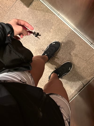 A person's legs and hands are visible in a close-up shot taken from a high angle. The person is wearing black Puma sneakers with white trim and laces, and black athletic shorts with a white marble-patterned waistband. They are holding a small set of scissors with a red handle and a black strap in their right hand. The background appears to be a textured, beige floor, possibly inside an elevator or a subway station, given the presence of a metallic wall on the right side of the frame. The lighting is artificial and somewhat dim. There is no discernible information to place this image in New York City, United States of America.