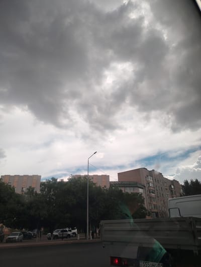 The media file depicts a cloudy sky over a street scene in Astana, Kazakhstan. The sky is filled with dark, heavy clouds, suggesting an impending storm or overcast weather. In the foreground, the back of a grey truck is visible, with part of its license plate showing the text "KZ 555OMG 01". Beyond the truck, several parked cars are visible on the side of the road, including a silver sedan and a white SUV. Trees line the street, and behind them, multi-story apartment buildings are present. One building appears to be under construction or renovation, with its facade featuring a pattern of lighter and darker bricks. The lighting is dim, consistent with a cloudy day. No people are clearly visible in the frame. The overall impression is of an urban environment on a day with challenging weather conditions.
