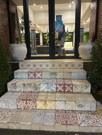 The image shows the tiled entrance steps of a building in Sukawati, Indonesia. The steps are adorned with a variety of colorful, patterned ceramic tiles, featuring floral and geometric motifs. On the left, a large, light-colored pot sits near a brick wall and lush green foliage. To the right, another large, light-colored pot is visible next to more greenery. The entrance doors are glass, revealing a well-lit interior with display cases and a person behind a counter. A "PUSH OPEN" sign is visible on the glass door, along with "JAM BUKA OPENING HOURS 9 AM - 9 PM". A person in a blue shirt and patterned shorts stands near the entrance, facing into the building. The ground in front of the steps is dark and appears to be asphalt. A single pink flower petal rests on one of the lower steps.
