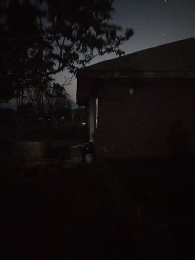 The image is a low-light photograph of an exterior scene at dusk or night. On the right side of the frame, a dark-colored building with a sloped roof dominates the view. A single, dimly lit window or opening is visible on the side of the building. To the left of the building, a tree with dark foliage is silhouetted against the sky. In the foreground, the ground is dark and appears to be unpaved. A round, concrete structure, possibly a well or a planter, is visible in the lower left portion of the image. Some objects are present in the darker areas of the scene, including what appears to be a bucket and possibly some other debris. The sky above is dark with a few faint stars visible. The overall impression is a rural or semi-rural setting in or near Iyara, Nigeria, characterized by its low illumination.