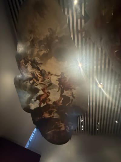 The image shows a ceiling with a painted mural in San Bernardino Tlaxcalancingo, Mexico. The mural depicts numerous human figures, possibly angels, in a cloud-like formation. The ceiling also has a modern striped design above the mural. A light source, likely a spotlight, shines directly onto the mural, creating a bright glare. Part of a dark-colored wall is visible in the lower right corner.