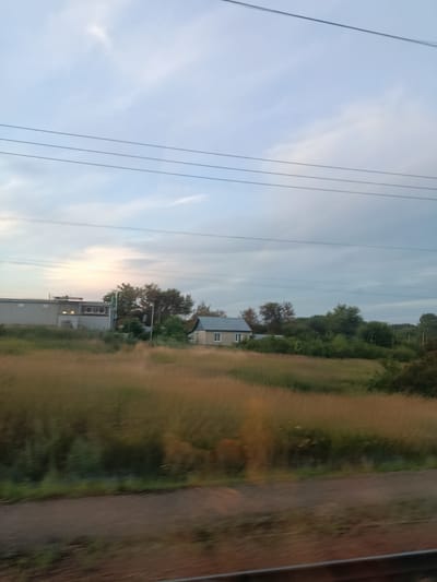 A photograph captured from a moving vehicle in Miloslavskoye, Russia, shows a rural landscape under a clouded sky. In the distance, a small, light-colored house with a grey roof is visible amidst a cluster of green trees. To the left of the house, a large, grey, industrial-looking building with circular vents is situated. The foreground is dominated by a field of tall, dry grass, suggesting late summer or autumn. Power lines stretch horizontally across the upper portion of the frame, and a blurred section of what appears to be a train track or road is visible at the bottom. The overall impression is one of a quiet, somewhat isolated rural setting.