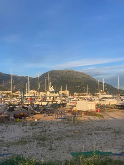 The image depicts a marina in the city of Bar, Montenegro, under a clear blue sky with sparse clouds. The foreground is a gravelly, unpaved area with scattered vegetation and some debris. A few vehicles, including two dark-colored cars and a green van, are parked near the boats.
The middle ground is dominated by a collection of boats of various sizes, many with tall masts, suggesting they are sailboats. Some boats are docked, while others appear to be undergoing maintenance or are stored on land. One prominent white motor yacht has "BARBARA" written on its side. A white truck with text and a red barrel are also visible.
In the background, buildings of the city of Bar are visible, and a large, rugged mountain range rises behind the city. The lighting suggests it is either early morning or late afternoon, with the sun casting long shadows. The overall scene conveys a working marina environment.