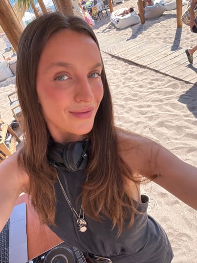 In Sutomore, Montenegro, a woman with long brown hair and headphones around her neck smiles directly at the camera. She is positioned in the foreground, with a wooden deck, sand, and beachgoers in the background. The woman is wearing a dark gray sleeveless top and a silver necklace with two pendants. A laptop and DJ equipment are visible on a wooden surface to her left. The scene appears to be a beachside setting, possibly a cafe or a DJ booth set up on the sand. The overall lighting suggests a sunny day.
