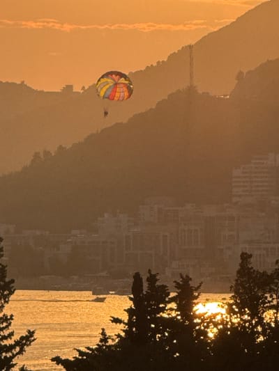 A parasailer is visible in the sky above a coastal city during sunset in Vrijesno, Montenegro. The parasail is colorful, with segments of red, yellow, blue, and green. Below the parasailer, the city is built on the slopes of a mountainous coastline. Buildings, some appearing to be hotels or apartment complexes, are clustered together. In the foreground, dark silhouettes of trees frame the scene. The water in front of the city reflects the warm orange and yellow hues of the sunset, with a few small boats visible on the surface. The overall atmosphere is serene and captures a moment of leisure activity against a picturesque backdrop.