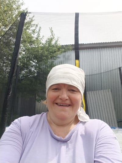 A medium shot shows a smiling woman wearing a white headscarf and a light purple T-shirt. She is positioned in front of a trampoline, which is surrounded by a black mesh net and has a gray, corrugated metal fence behind it. Green trees are visible through the mesh. The location is Красный Маяк, Russia.