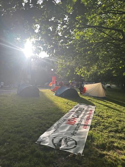 This image depicts an outdoor scene in a public green space, likely a park, illuminated by the late afternoon or early evening sun which casts long shadows and produces a significant lens flare in the upper left. The setting is identified by a prominent banner as the La Calzada neighborhood in Gijón/Xixón, Spain.

The foreground features a wide expanse of neatly maintained grass, with several large deciduous trees lining the left and right sides, their foliage creating a dappled light effect. A large white banner with black and red text lies flat on the grass in the lower foreground, held down by small rocks. The banner reads "NO A LA CONTAMINACIÓN / NO AL RUIDO / NO A LAS MERMAS" (NO TO POLLUTION / NO TO NOISE / NO TO LOSSES), followed by "NO + CAMIONES" (NO MORE TRUCKS) in large red letters, and then "Por un barrio libre de transporte pesado y ruidoso." (For a neighborhood free of heavy and noisy transport.) The banner concludes with "A.VV. DE LA CALZADA - 'AL FONDO CAMÍN'", identifying the "Neighborhood Association of La Calzada." A graphic of a truck with a red circle-and-slash symbol is also visible on the banner.

In the midground, several camping tents are erected on the grass, indicating an encampment. From left to right: a dark grey pop-up tent is centrally placed, slightly obscured by shadows. Further back and to its right, a smaller orange tent is partially visible. To the right of the grey tent, a larger, light beige/off-white traditional tent with blue and orange trim is set up. A person is partially visible near the entrance of this beige tent, bending down. Another dark green pop-up tent is visible on the far right. More tents are visible further down the grassy area in the distance, suggesting a larger gathering.

In the background, beyond the immediate grassy area and a paved path, several multi-story buildings with traditional facades are visible, characteristic of urban European architecture. A street lamp stands between the trees and the buildings. Scattered individuals and small groups of people are visible in the background, some near a red and white structure that might be another tent or stand. The overall scene suggests a sustained protest or community gathering.
