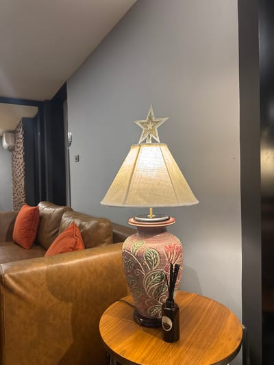 In a contemporary living room setting, likely in Bangkok, Thailand, a warmly lit table lamp with a star ornament sits on a small, round wooden table. The lamp's shade is a neutral beige, and its base is a decorative ceramic vase featuring a floral motif in shades of pink, green, and red. Next to the lamp on the table is a small dark glass bottle with diffuser reeds, suggesting a home fragrance. Behind the table, a plush, brown leather sofa with two orange accent pillows is visible. The room's walls are painted in a cool grey, and a black vertical column is present on the right side of the frame. In the background, another part of the room shows an air conditioning unit mounted on a wall with a textured, mosaic-like pattern. No people are present in the scene.