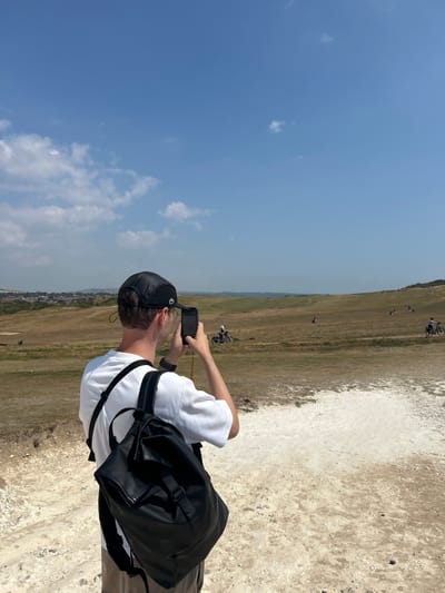 A man wearing a white t-shirt and a black baseball cap is standing and taking a photo with his phone in a landscape with dry grass under a clear blue sky with some clouds. He has a black backpack on. In the background, there are several people on bicycles and a town in the distance. The foreground consists of dry, sandy ground with some rocks.