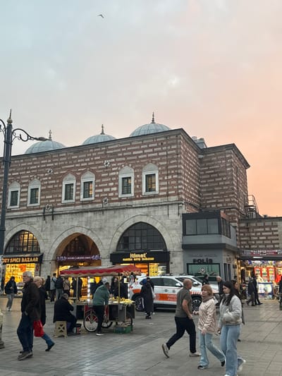 The image depicts a bustling pedestrian scene at dusk in front of the Spice Bazaar (Misir Çarşısı) in the Eminönü quarter of the Fatih district, Istanbul, Turkey. The sky exhibits an overcast, fading light, with grey and light blue hues transitioning to a warm orange-pink towards the horizon. A single bird is visible in the upper left sky.
The prominent structure is a multi-story historical building, identified by signs as "MISIR ÇARŞISI (1664) SPICE BAZAAR." Its lower level features large stone arches housing storefronts, including "KOZY SPICE BAZAAR" and "Itimat PABUC SANAYI VE TICARET." The upper facade is constructed with alternating courses of red brick and light-colored stone, punctuated by several arched windows with dark frames. The roofline is characterized by three prominent, light-colored domes, each topped with a finial. To the immediate right of the historical building, a modern, two-story annex with dark cladding houses a "POLIS" (Police) station, with two white "TURIZM POLISI" vehicles parked directly in front.
On the paved pedestrian area in the foreground and midground, approximately two dozen individuals are present. Most are adults, casually dressed, engaged in walking or standing. In the center midground, a red-canopied street vendor cart, possibly selling roasted corn or chestnuts, is attended by several people, including a man in a green hoodie. Another man sits on a stool next to the cart. A decorative black metal streetlamp with ornate scrollwork stands on the far left. Individuals move in various directions across the stone-paved ground.