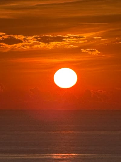 A serene sunset over the ocean creates a vibrant canvas of orange and red hues. The sun, a bright white orb, is positioned slightly left of center, casting a warm glow across the scene. Wispy clouds are scattered across the sky, some tinged with the fiery colors of the sunset. The ocean below reflects the sky's radiance, with subtle horizontal lines indicating gentle waves. The horizon is a sharp, clean line separating the sky from the sea. The overall atmosphere is one of tranquility and natural beauty. This image could be captured on Lý Sơn Island, a volcanic island in Quảng Ngãi Province, Vietnam, known for its stunning coastal scenery and dramatic sunsets.